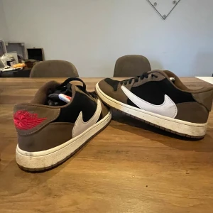 Nike Air Jordan 1 Low Travis Scott - Säljer ett par Nike Air Jordan 1 Low Travis Scott med svart och brunt mocka, vit swoosh och röda detaljer på hälen. Skorna har klassisk snörning och en platt sula. Perfekta för dig som gillar streetwear och vill sticka ut med en ikonisk sneaker.