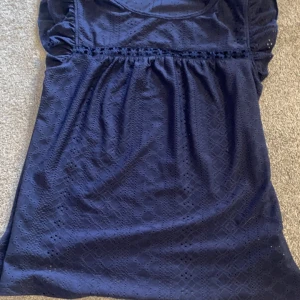 Mörkblå blus med spetsdetaljer - Säljer en mörkblå blus med fina spetsdetaljer och diskret mönster. Blusen har korta ärmar med volang och rund halsringning. Perfekt för dig som gillar en söt och stilren look.