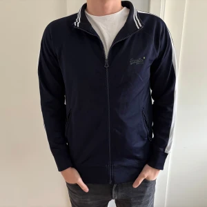 Mörkblå ziptröja från Superdry - Sportig mörkblå ziptröja från Superdry med vita ränder på kragen och längs ärmarna. Tröjan har dragkedja framtill, två fickor med zip och diskret logga på bröstet. 
