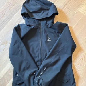 Svart vindjacka från Haglöfs - Svart vindjacka från Haglöfs med huva och hel dragkedja framtill. Jackan har meshfoder på insidan och diskret logga på bröstet. Perfekt för blåsiga dagar och outdoor-äventyr. Snygg och funktionell design som passar till det mesta.