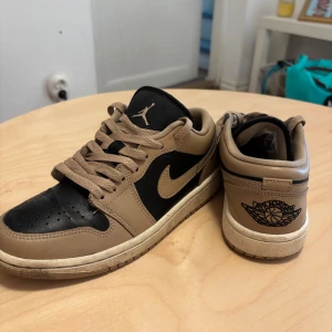 Nike Air Jordan 1 Low Desert - Säljer ett par Nike Air Jordan 1 Low i färgen Desert. Skorna har en snygg kombination av beige och svart skinn, klassisk snörning och den ikoniska Air Jordan-loggan på hälen. Perfekta för dig som vill ha en stilren och trendig sneaker med låg profil.