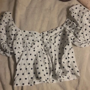 Vit prickig blus med puffärmar - Superfin vit blus med svarta prickar och puffiga korta ärmar. Söt och trendig modell med markerad midja och knappar framtill. Perfekt för dig som gillar en lekfull och feminin stil. Gjord i ett lätt och luftigt bomullstyg.