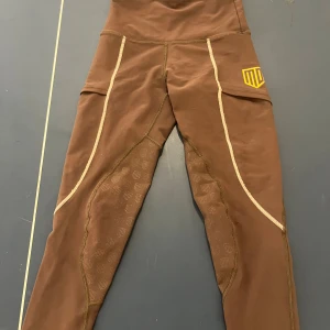 Bruna ridleggings från Maya Delorez - Snygga bruna ridleggings från Maya Delorez med hög midja och smarta fickor på sidorna. Silikonprint för extra grepp på insidan av benen och kontrastsömmar i beige. Perfekta för ridning och riktigt bekväma med stretchigt material.