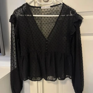 Svart prickig blus från Pull&Bear - Säljer en svart transparent blus från Pull&Bear med prickigt mönster, v-ringning och knappar framtill. Blusen har långa ärmar med volangdetaljer på axlarna och smock vid ärmsluten. Perfekt att styla med jeans eller kjol.