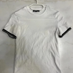 Vit t-shirt med pärldetaljer från Zara - Vit t-shirt från Zara i bomull med korta ärmar. Ärmsluten har svarta spetsdetaljer och pärlor som ger en unik och cool touch. Perfekt för dig som vill ha något enkelt men ändå med en twist.