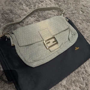 Fendi Baguette axelväska monogram - Säljer en ikonisk Fendi Baguette vintage axelväska i beige med klassiskt Fendi-monogram över hela väskan. Väskan har ett snyggt spänne framtill i metall och justerbar axelrem. Materialet är canvas med detaljer i skinn och insida med dragkedjefack. 