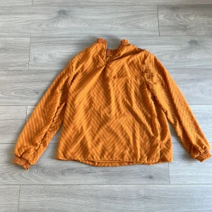 Orange långärmad blus med struktur - Säljer en orange långärmad blus med snyggt strukturmönster i sicksack. Blusen har smockad hög hals och smockade ärmslut för en cool och bekväm look. Materialet känns lätt och har en subtil glans. Perfekt för dig som vill sticka ut med färg och mönster.