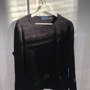Svart långärmad tröja från Polo Ralph Lauren - Svart långärmad tröja från Polo Ralph Lauren med rund halsringning och diskret blå logga broderad på bröstet. Klassisk och stilren modell som passar perfekt till jeans eller chinos. Sitter som M/L