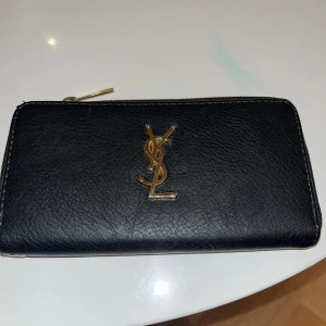 Svart YSL plånbok i skinn - Snygg svart plånbok från YSL i äkta skinn med guldigt YSL-logo framtill. Plånboken har dragkedja runtom, flera kortfack och ett myntfack med dragkedja. Perfekt för dig som vill ha en stilren och lyxig accessoar i väskan.