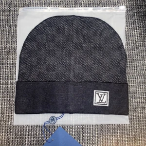 Svart Louis Vuitton mössa med logga - Snygg svart mössa från Louis Vuitton med diskret rutigt mönster och uppvikt kant. På framsidan sitter en vit patch med LV-loggan. Mössan är tillverkad i mjukt material som känns lyxigt och passar perfekt för att höja din streetstyle.