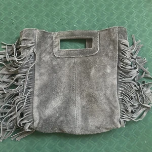 Grå mockaväska med fransar - ”Maje väska” liknande Snygg grå handväska i mocka med rektangulärt handtag och coola fransar längs sidorna. Perfekt accessoar för dig som vill sticka ut med en bohemisk touch. Väskan har en mjuk känsla och unik design. Äkta mocka🤎
