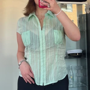 Mintgrön randig skjorta från H&M - Snygg mintgrön skjorta från H&M med tunna vertikala ränder och två bröstfickor. Skjortan har korta ärmar, klassisk krage och knäppning framtill. Perfekt för en fräsch och luftig look under varmare dagar. Axelbredd: 45cm. Längd: 64cm. Byst: 52cm.