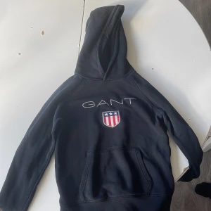 Svart hoodie från GANT med logga - Svart hoodie från GANT med broderad logga och amerikanskt sköldmärke på bröstet. Klassisk känguruficka framtill och stor huva. Perfekt för dig som gillar sportig och stilren streetwear.
