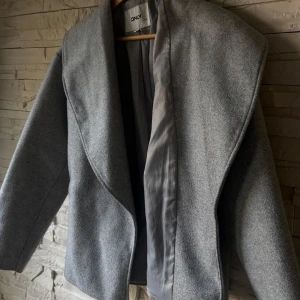 Grå kappa från ONLY - Stilren grå kappa från ONLY. Jackan har en rak passform och är perfekt för dig som gillar minimalistisk och clean stil. Materialet känns mjukt och har en ull-liknande känsla. Snygg att slänga över vilken outfit som helst. Jättebra inför hösten!