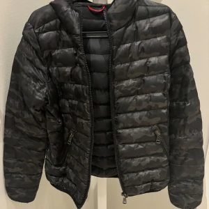 Svart camo pufferjacka från Moncler - Snygg svart pufferjacka från Moncler med camouflagemönster. Jackan har huva, två dragkedjefickor och klassisk Moncler-logga på ärmen. Perfekt för dig som vill ha en streetig och trendig look under kyliga dagar.