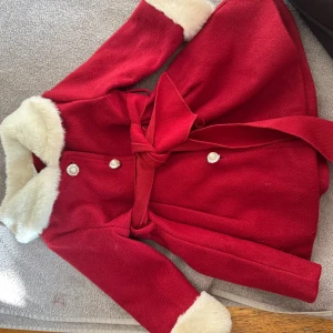 Röd kappa med fuskpäls och pärlknappar - Röd kappa med vit fuskpäls på krage och ärmslut, pärlknappar framtill och knytskärp i midjan. Kappan har en klassisk, lite utsvängd form och långa ärmar. Perfekt för vintern och julens alla tillfällen.