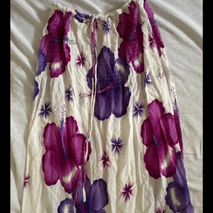 Vit långkjol med lila och rosa blommor - Superfin långkjol med stora blommor i lila och rosa på vit botten. Kjolen har snörning i midjan och är gjord i ett lätt och luftigt material, perfekt för varma dagar. Den har en avslappnad passform och ger en härlig sommarkänsla.