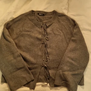 Grå kofta med snörning från Gina Tricot - Grå kofta från Gina Tricot i storlek XS. Koftan har lång ärm och snygga snörningsdetaljer framtill istället för knappar. Den är stickad och har en klassisk rund halsringning. Perfekt för lager-på-lager under hösten och vintern. Använd en gång