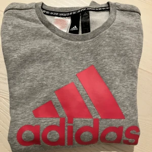 Grå Adidas sweatshirt  - Grå sweatshirt från Adidas med stor rosa logga och text framtill. Tröjan har rund hals, långa ärmar och ribbade muddar vid ärmslut och nederkant. Perfekt för dig som gillar sportig och avslappnad stil. 152cm 