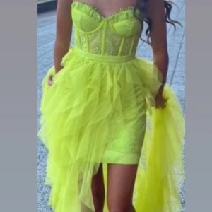 Neongul volangklänning med spetsliv - Supercool långklänning i neongult med volanger i flera lager och spetsdetaljer på livet. Klänningen har en figurnära passform upptill, markerad byst och axelbandslös design. Materialet är lätt och skirt, perfekt för att sticka ut på bal eller fest.