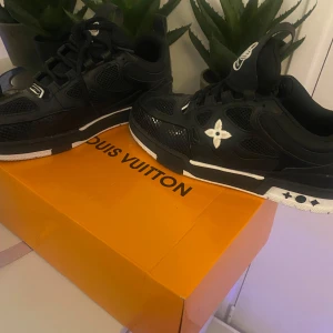 Louis Vuitton svarta sneakers - Snygga svarta Louis Vuitton sneakers med vita detaljer och ikonisk blomlogga på sidan. Skorna har meshpaneler för extra andningsförmåga, svarta snören och en vit sula med svarta mönster. Perfekta för dig som vill sticka ut med lyxig streetstyle.