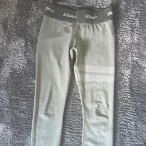 Stronger träningsbyxor - Snygga leggings från Stronger med bred resår i midjan där loggan STRONGER syns runtom. Byxorna har två vita ränder på ena benet och är tillverkade i ett stretchigt material som passar perfekt till träning. Dom är inte alls använda och är som nya!