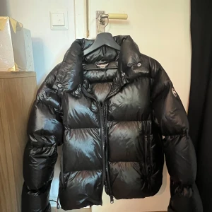 Svart pufferjacka från Colmar - Säljer en svart pufferjacka från Colmar med glansig finish och markerad krage. Jackan har lång ärm, dragkedja framtill och logotyp på ärmen. Passformen är loose och den är perfekt för kalla dagar. Fylld med dun och har justerbar nederkant med snörning. Storlek S men passar även M
