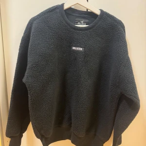 Svart fleece-tröja från Hollister - Mysig svart fleece-tröja från Hollister med broderad logga på bröstet. Tröjan har rund halsringning, ribbade muddar och är perfekt för kyliga dagar. Oversized passform för en avslappnad stil.