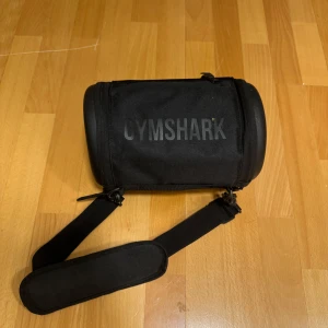 Svart axelväska från Gymshark - Säljer en svart axelväska från Gymshark med justerbar axelrem och hårda gavlar. Väskan är kompakt och perfekt för gymmet eller vardagen, med tydlig logga på framsidan. Tillverkad i slitstark polyester.