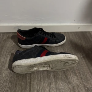 Gucci sneakers med metallic-detaljer - Snygga Gucci sneakers i mörkblått med GG-monogram, röd och blå rand på sidan samt metallic-detaljer i koppar och lila på hälen. Skorna har vit sula med unikt mönster undertill och klassisk snörning. Perfekta för dig som vill sticka ut med lyxig streetstyle.