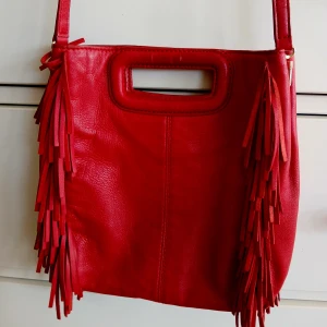 Röd fransad axelväska från Maje - Cool röd axelväska i skinn från Maje med fransar längs sidorna och både axelrem och handtag. Väskan har en fyrkantig form och stilren design med ett litet guldfärgat Maje-tryck vid handtaget. Perfekt för dig som vill sticka ut med en färgklick.