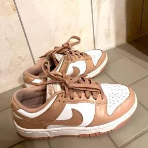 Nike Dunk Low i beige/rosa och vitt - Snygga Nike Dunk Low sneakers i beige/rosa och vitt med klassisk swoosh på sidan. Skorna har snörning, platt sula och perforerad tå för extra ventilation. Perfekta för dig som vill ha en stilren och trendig look till vardags.