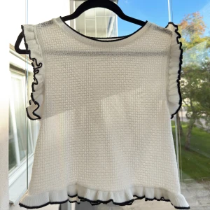 Vit stickad topp med volangdetaljer från Zara - Superfin vit stickad topp från Zara med volangkant på ärmar och nederdel, samt svarta kontrastsömmar. Toppen är ärmlös och croppad, med en lätt strukturerad yta. Aldrig använd men saknar prislapp