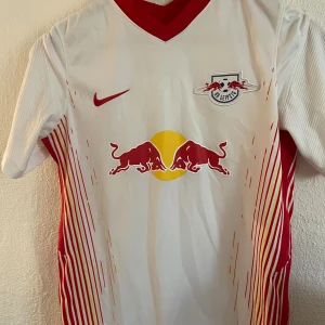 RB Leipzig vit matchtröja Nike - Snygg RB Leipzig fotbollströja från Nike i vitt med röda och gula ränder på sidorna. Tröjan har klubbmärke och Red Bull-logga på bröstet samt röd V-ringning. Tillverkad i lätt och ventilerande material, perfekt för träning eller match.