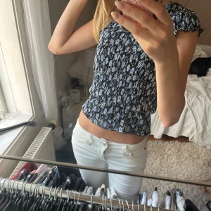 Blommig blå och svart blus med volangärm - Supersöt blus med blått och vitt blommönster på svart botten. Toppen har små volangärmar och smockad, figurnära passform som ger en snygg siluett. Perfekt till jeans eller kjol för en fräsch look. Materialet känns lätt och luftigt.