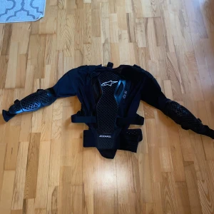 Svart skyddsjacka Alpinestars M - Säljer en svart Bionic Action V2 Protection Jacket från Alpinestars. Jackan har robusta skyddsplattor på bröst, rygg, axlar och armar med bikakemönster och justerbara remmar i midjan. Materialet är polyester och polyuretan, perfekt för dig som kör motocross eller downhill.