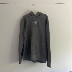 Grå Jordan hoodie med tryck - Snygg grå hoodie från Jordan med tryck på bröstet och ryggen där det står 'PARIS'. Tröjan har huva, långa ärmar och en dragkedjeficka på sidan. Perfekt för dig som gillar streetwear och sportig stil. Tröjan är även kopt i PSG shop i Paris.