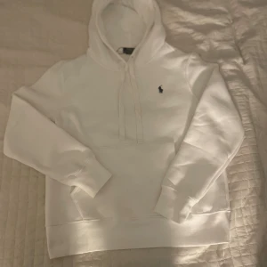 Vit hoodie från Polo Ralph Lauren - Vit hoodie från Polo Ralph Lauren med klassisk blå logga på bröstet. Tröjan har huva med snörning, stor magficka och ribbade muddar vid ärmslut och nederkant. Mjuk bomullskvalitet och clean look som alltid funkar. Skriv för mer info 