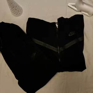 Svarta Nike mjukisbyxor med snörning - En är S och en är XS Svarta mjukisbyxor från Nike med snyggt tryckt logga på benet och praktisk snörning i midjan. Byxorna har en relaxed passform och är tillverkade i mjukt material som är perfekt för chill eller träning. Snygga detaljer med dragkedja på benet.