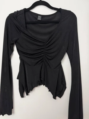 Svart långärmad topp från SHEIN - Svart långärmad topp från SHEIN med v-ringning och snygg rynkdetalj framtill. Toppen har asymmetrisk nederkant och är tillverkad i ett lätt, stretchigt material som ger en slim passform. Perfekt för dig som gillar trendiga och unika plagg.