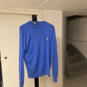 Blå tröja från Polo Ralph Lauren! - Slim fit blå tröja från Polo Ralph Lauren med klassisk rund hals och den ikoniska gula broderade loggan på bröstet. Långärmad och tillverkad i mjukt merinoull som passar perfekt till jeans. Passar dig runt 172 till 182 cm lång!