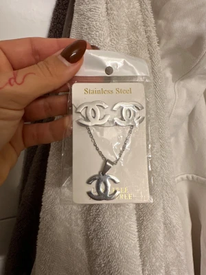 Set med halsband och örhängen - Säljer ett matchande set med halsband och örhängen i rostfritt stål, båda med den ikoniska CC-loggan. Smyckena är silverfärgade och har en stilren, modern look som passar perfekt till din outfit.