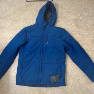 Blå pufferjacka Peak Performance - Blå pufferjacka från Peak Performance med huva och dragkedja framtill. Jackan har quiltad design, långärmade ärmar och praktiska fickor. Perfekt för kyliga dagar och har en clean, sportig look. Materialet är polyester och jackan är i mediumstorlek.