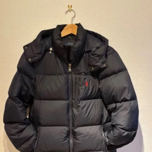 Svart pufferjacka från Polo Ralph Lauren - Svart pufferjacka från Polo Ralph Lauren med ikonisk röd logga på bröstet. Jackan har hög krage, huva och tvåvägsdragkedja med RL-detaljer. Fylld med dun och har en quiltad design som ger extra volym och värme. Perfekt för kalla dagar.