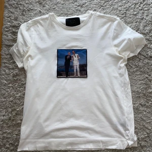 Vit T-shirt Limitato med print - Vit T-shirt från Limitato med ett stort, färgstarkt print på bröstet som föreställer två personer i kostym. Klassisk rund hals och korta ärmar. Materialet är mjuk bomull och t-shirten kommer i exklusiv originalförpackning med Limitato-logga.