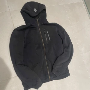 Svart hoodiejacka från Sail Racing - Svart hoodiejacka från Sail Racing med dragkedja framtill och bröstficka med logotyp. Jackan har huva med tryckt emblem och ribbade muddar vid ärmslut och nederkant. Perfekt för chill dagar och streetstyle.