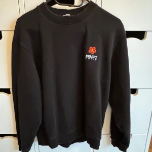 Svart sweatshirt från Kenzo XS (14-16 years) - Svart sweatshirt från Kenzo i storlek XS med rund hals och ribbade muddar. På bröstet finns en röd broderad blomma och Kenzo Paris-logga i vitt. Tröjan har en klassisk, loose passform och är tillverkad i mjuk bomull som är skön att bära.