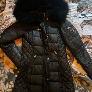 Svart lång Rockandblue dunjacka - Svart lång dunjacka från Rockandblue med quiltad design, stor fluffig fuskpäls på huvan och flera dragkedjor i guld. Jackan har figurnära passform, snygga detaljer på ärmarna och Rockandblue-logga på ena ärmen. Perfekt för kalla dagar.