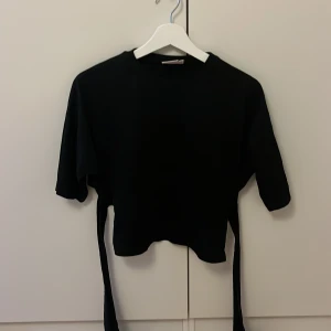 Svart topp med knyt från Nelly XS - Svart croppad topp från Nelly i storlek XS. Toppen har halvlånga ärmar och långa band som knyts i midjan för en snygg omlott-effekt. Enkel och stilren design som passar till många olika looks.