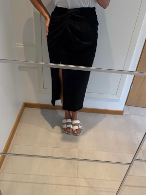 Svart omlottkjol med slits från H&M - Svart midikjol med omlottkänsla och knutdetalj i midjan från H&M. Kjolen har en snygg slits framtill och ett diskret strukturmönster i tyget. Tillverkad i viskos, polyester och polyamid för en lätt och följsam känsla. Svart midikjol med mönstrat prickigt tyg, draperad dekorativ knut och slits framtill. Perfekt till julfest, nyår, osv.